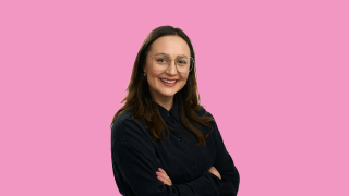 VAKEn psykologin Sonja poseeraa mustassa paidassa vaaleanpunaista taustaa vasten, hymy kasvoillaan.