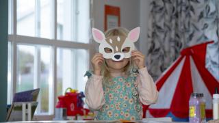 Leikki-ikäinen lapsi seisoo pöydän ääressä ja pitää eläinnaamaria kasvoillaan.