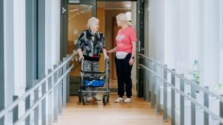 Vanhus rollaattorilla juttelee käytävällä hoitajan kanssa.