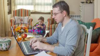 Henkilö työskentelee tietokoneella kotona, taustalla lapsi leikkii